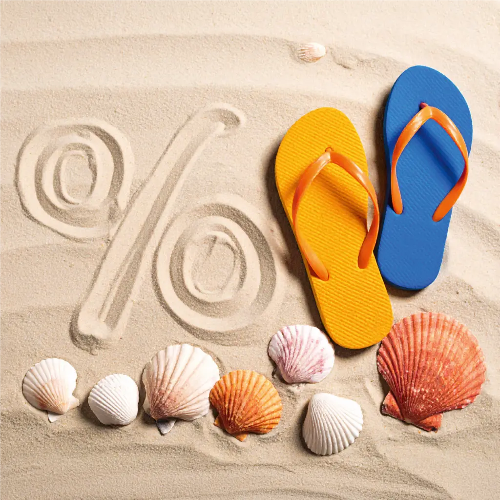Ein in den Sand gezeichnetes Prozentzeichen. Daneben liegen zwei Flip-Flops sowie einige Muscheln im Hand.